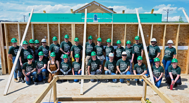 A group of 30 Ovintiv volunteers wearing green hard hats and matching “Where Industry Builds Impact” t-shirts pose in front of a framed wall at the Habitat Industry Build site in Airdrie.