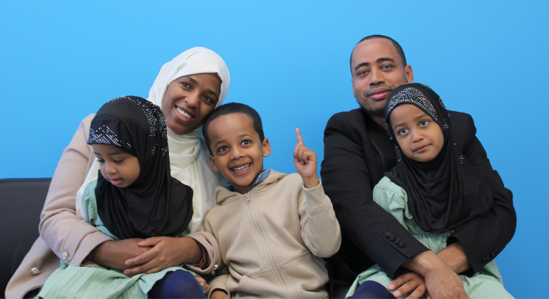 A family of five sits in front of a blue wall. The mom is smiling big, holding 2 small children and the father smiles with a daughter on his lap. 