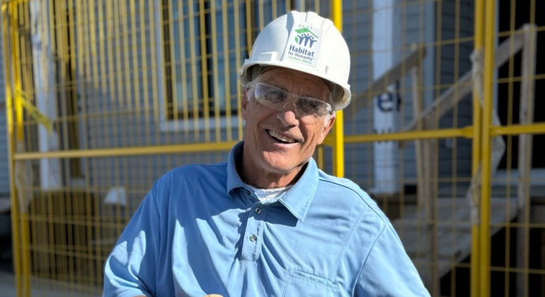 Volunteer Crew Leader Jeff wearing a white hardhat and a blue button up shirt takes a break to sit in front of a Habitat build in Calgary.