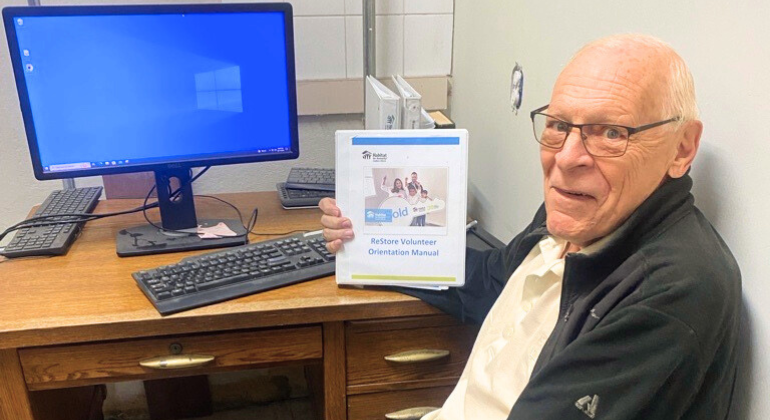 Doug sits at the computer proudly showing the ReStore Volunteer Orientation Manual. 