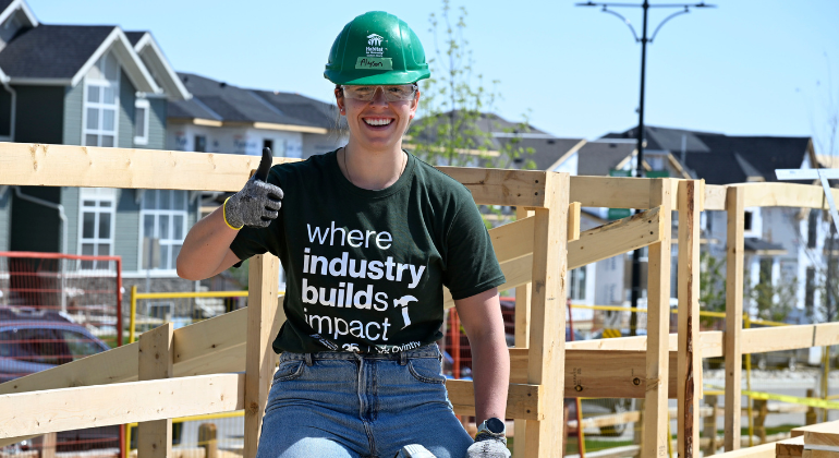 An Ovintiv volunteer wearing a green Habitat hard hat and “Where Industry Builds Impact” t-shirt smiles and gives a thumbs up while seated on the build site, surrounded by partially framed wooden walls..