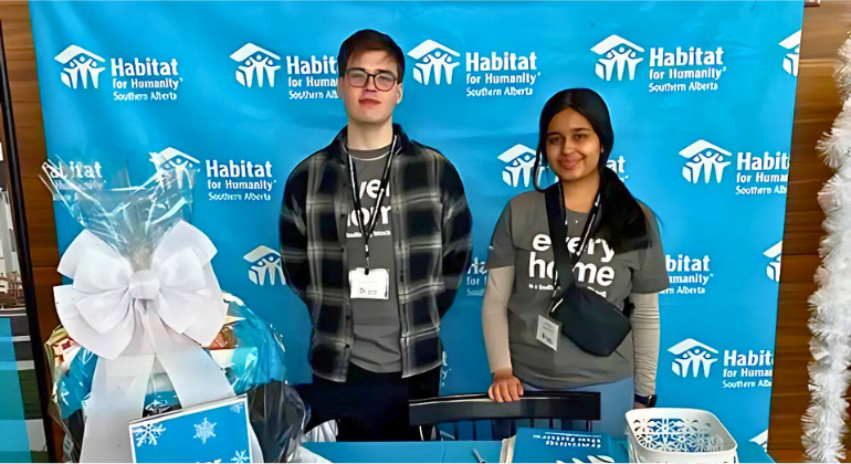 Two UCalgary students standing in front of a Habitat for Humanity banner at a community booth.