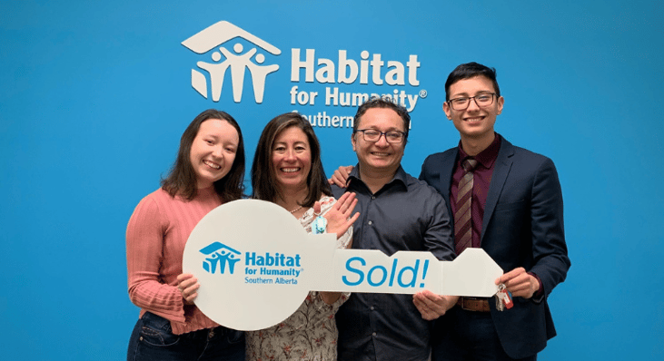 A family holding a large Habitat cardboard key with "sold!" in front of a blue wall with the Habitat for Humanity logo behind them