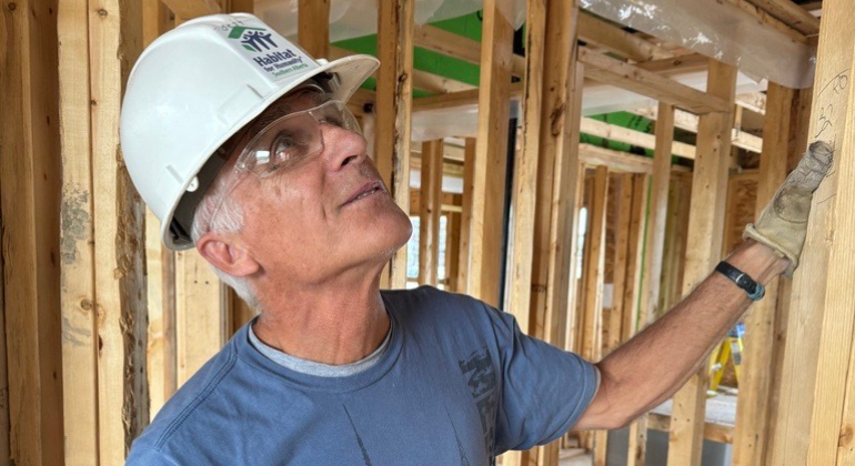 A man wearing a hard hat and safety glasses looking at a section of studs in a home under construction.