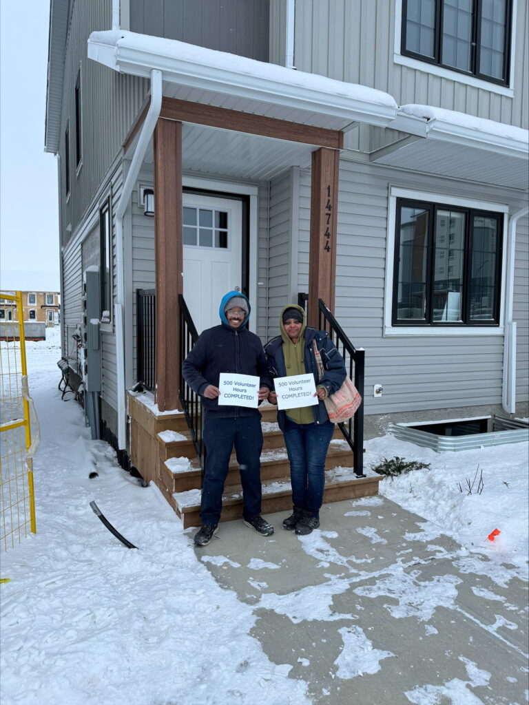 A couple holding paper which reads "500 volunteer hours completed!" in front of a Habitat townhome