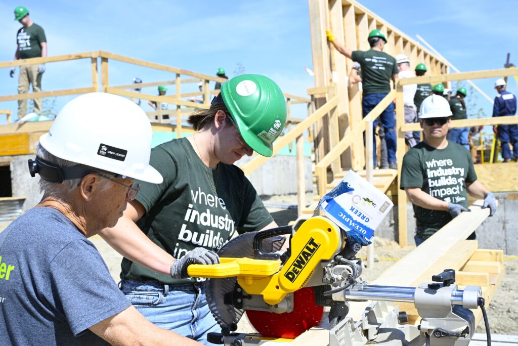 A volunteer wearing a green hard hat and “where industry builds impact” t-shirt uses a power saw under the guidance of a Habitat for Humanity crew leader. Other volunteers are working on framing in the background at a sunny build site.