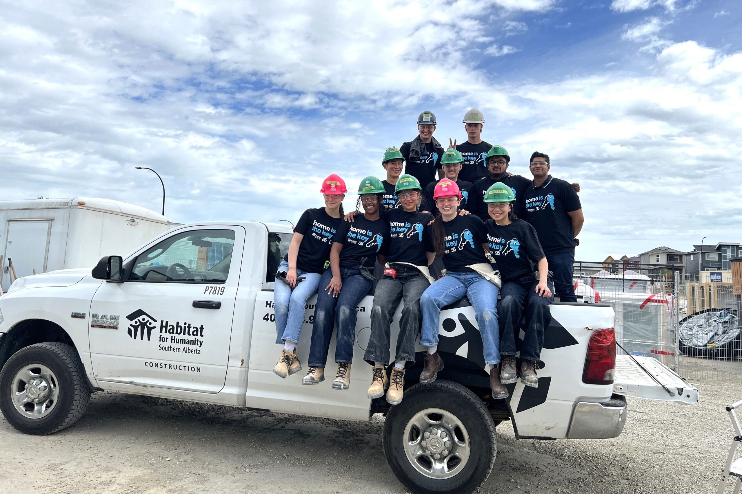 A group of Habitat for Humanity Southern Alberta volunteers wearing hard hats and matching “Home is the Key” t-shirts sit and stand on a white Habitat construction truck at a build site, smiling at the camera under a partly cloudy sky.