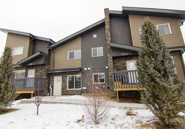 A row of modern townhomes with a mix of beige, brown, and dark grey siding, featuring stone accents and small front porches. Snow covers the ground and trees are planted in front of the homes, suggesting a winter season.