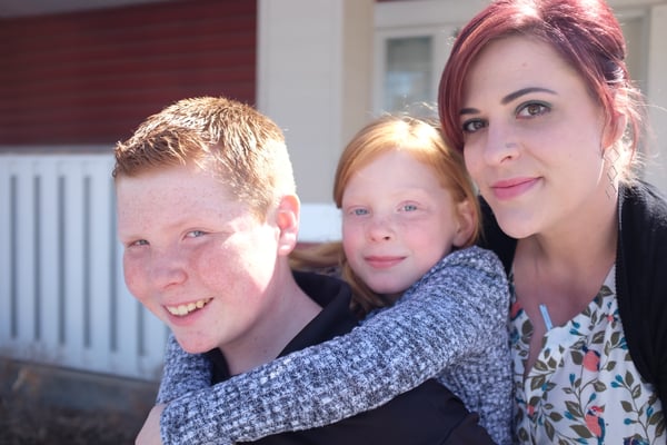 A smiling woman with red hair poses outside with two children, a boy and a girl. The girl, with long red hair, has her arms around the boy's shoulders in a piggyback-style hug. They are standing in front of a house with white and red siding, enjoying a sunny day.