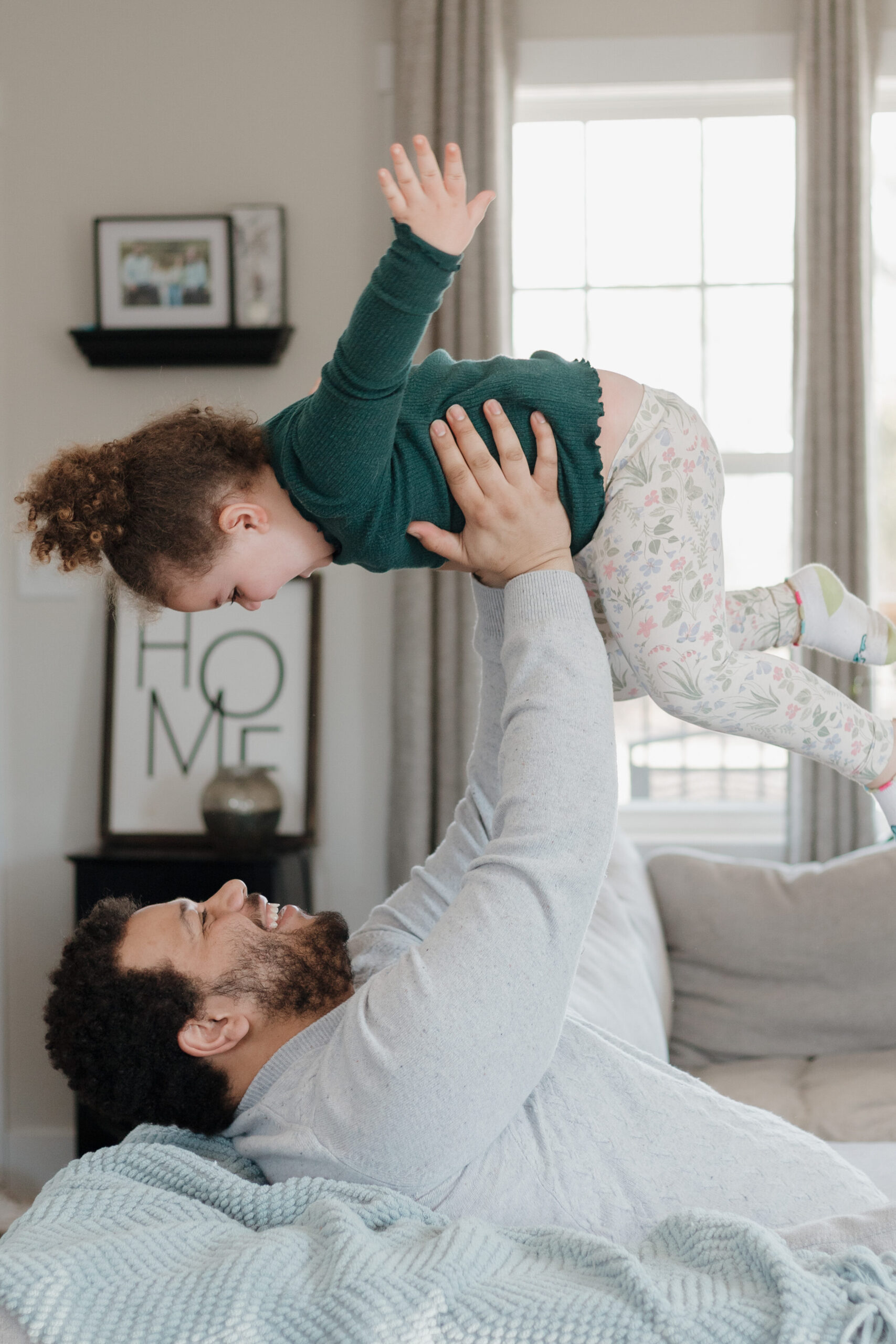 a Dad playfully tosses his little girl above his head while they play on the couch.