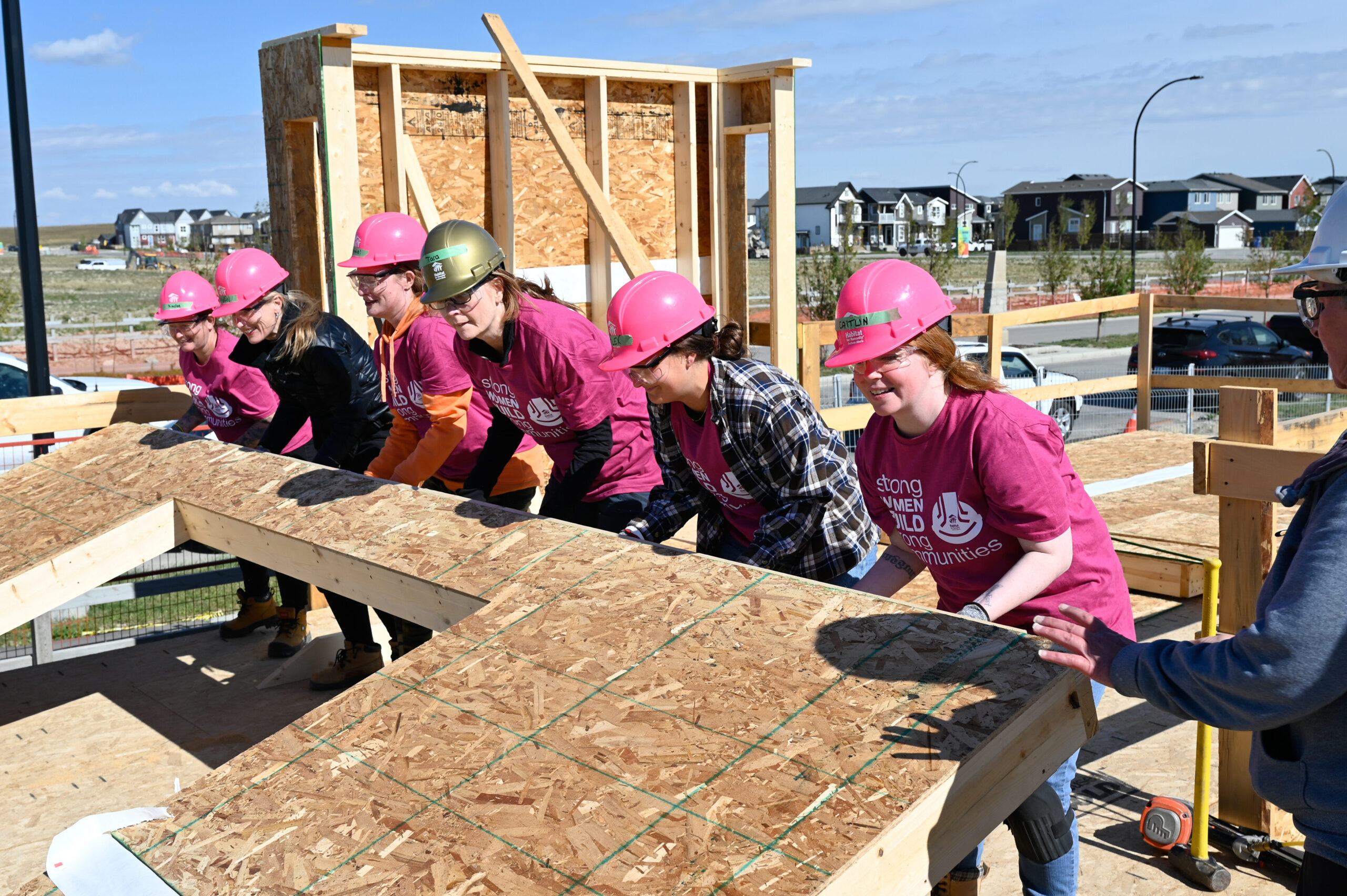 A group of Women Build volunteers wearing pink shirts and hard hats work together to lift a framed exterior wall section into place at a Habitat for Humanity build site, guided by a crew leader.