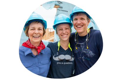 Three Habitat volunteers wearing blue hardhats smiling on a construction site.