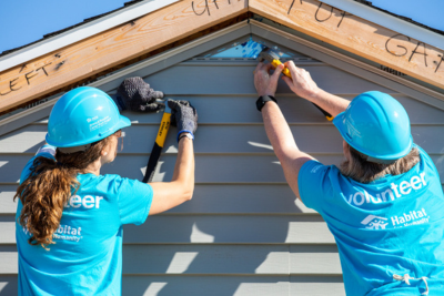Two volunteers wearing Habitat blue volunteer shirts and hardhats work together to install siding.