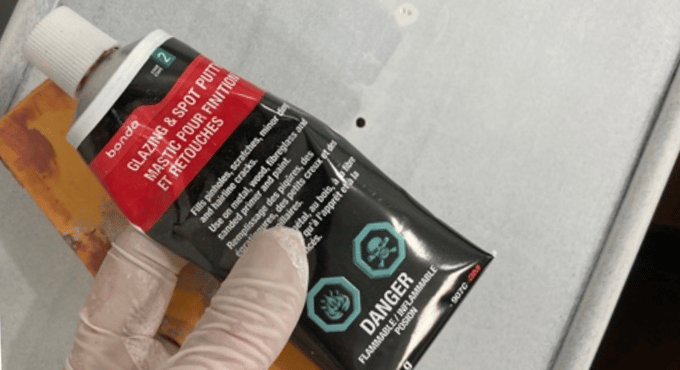 a closeup of a hand wearing a latex glove, holding a small tube of Bondo repair putty