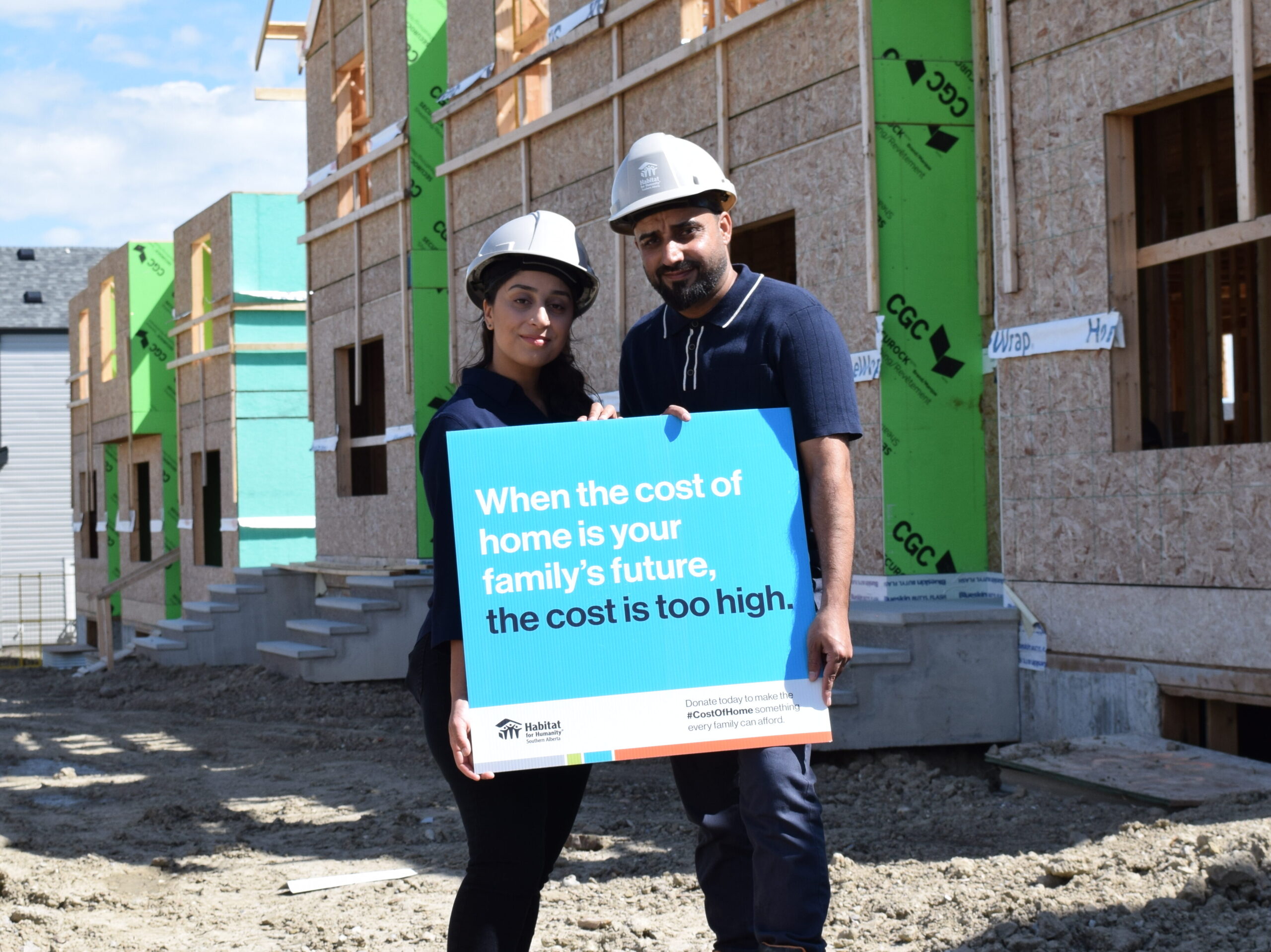 Iqra and Muhammad stand in front of their unfinished Habitat home holding a sign that says "When a home costs half your paycheque the cost is too high".