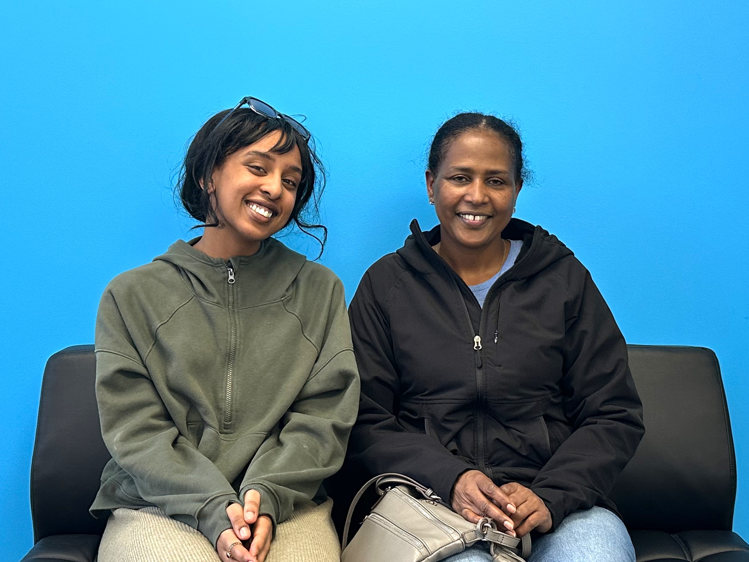 Dama and Chaltu sit on a black couch in front a bright blue wall.