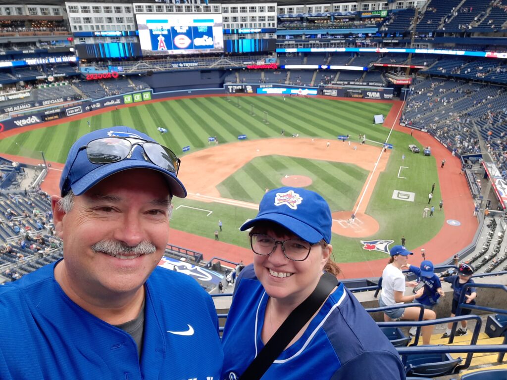 Howard and his wife smiling for a selfie in the stands at Rogers Centre, wearing Blue Jays gear with the baseball field and scoreboard visible in the background.