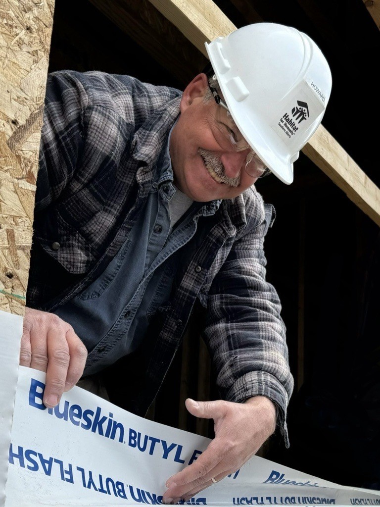 Howard smiling while installing Blueskin flashing on a Habitat build site, wearing a white hard hat labeled with his name and a plaid jacket.