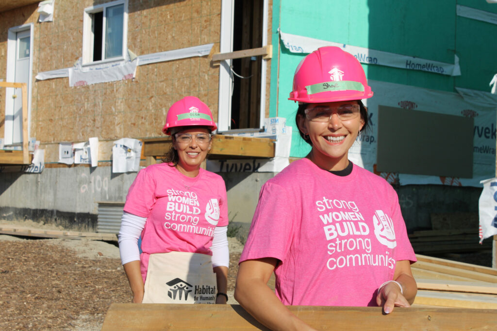 Two women wearing hardhats carrying a piece of wood