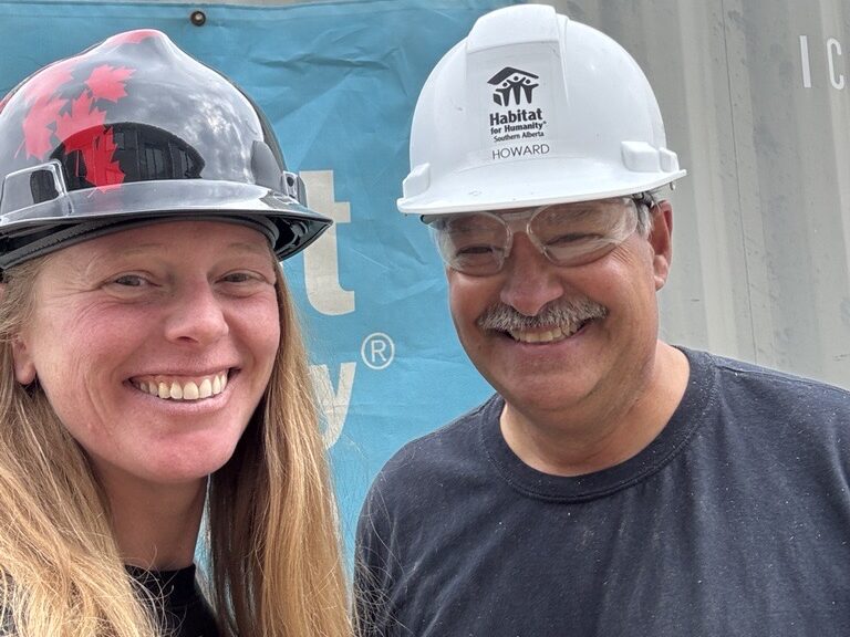 Howard and Trish smiling in their hard hats on the Habitat build site, standing in front of a blue sign and seacan.