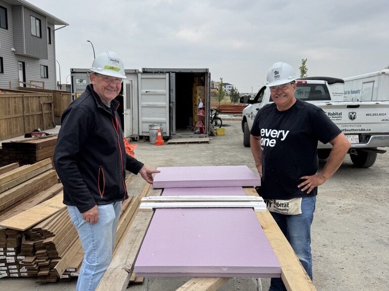 Two Habitat volunteers, Howard and James, smiling while measuring purple insulation panels on a build site with lumber, trucks, and construction materials in the background.