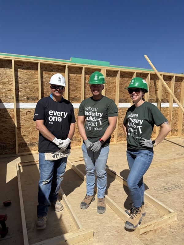 Howard standing on a Habitat build site with two volunteers wearing hard hats and safety gloves, smiling in front of a framed wall under a clear blue sky.