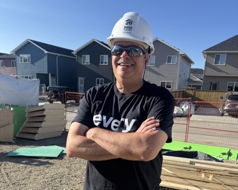 Howard stands with arms crossed on a Habitat build site wearing a white hard hat.