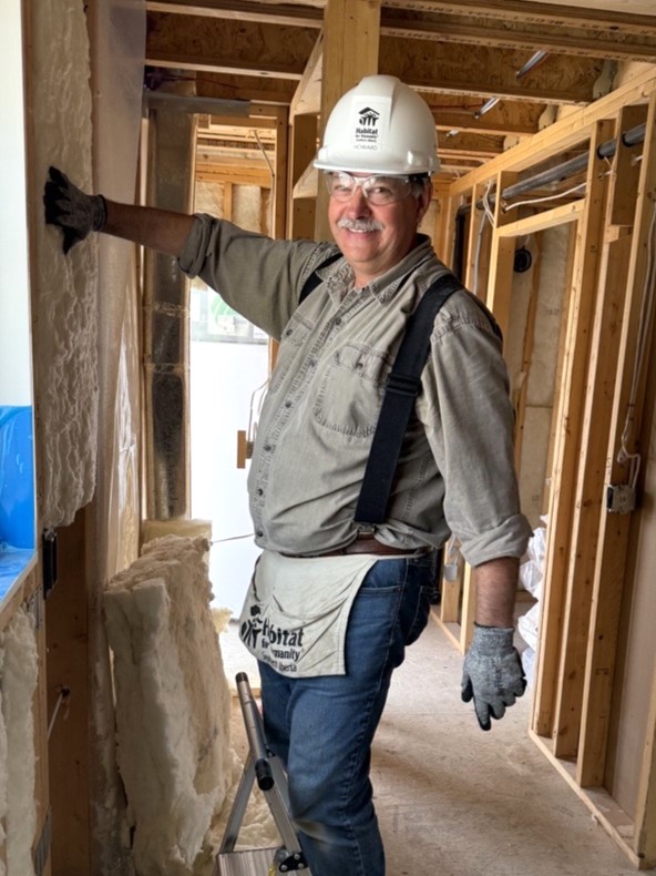 Howard smiling while placing insulation between wall studs on a Habitat build site, wearing a white hard hat, gloves, and a Habitat tool belt.