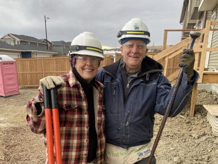 Two volunteers wearing hardhats and PPE stand smiling on the construction site in Airdrie.