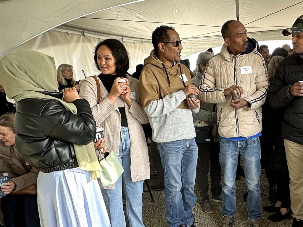 Addis and Eyob stand among a crowd under a white tent chatting with their future neighbours. 
