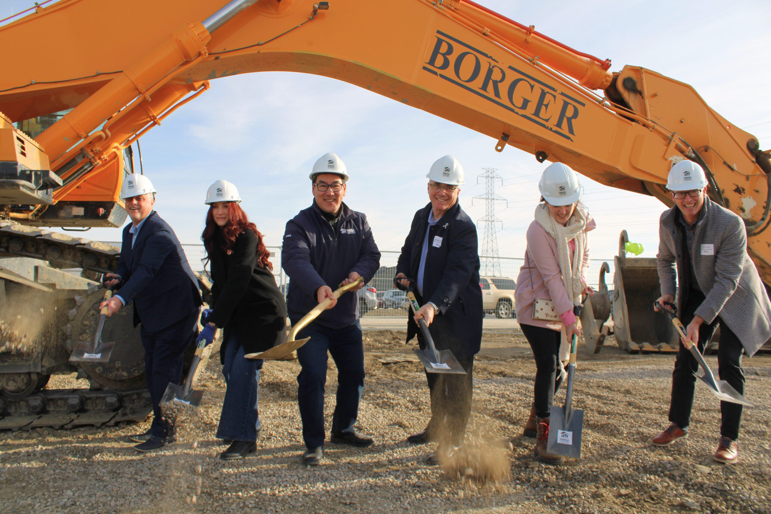 Five men and women make a cermonial groundbreaking scoop with shovels in white hardhats standing on dirt with an excavator in the background.