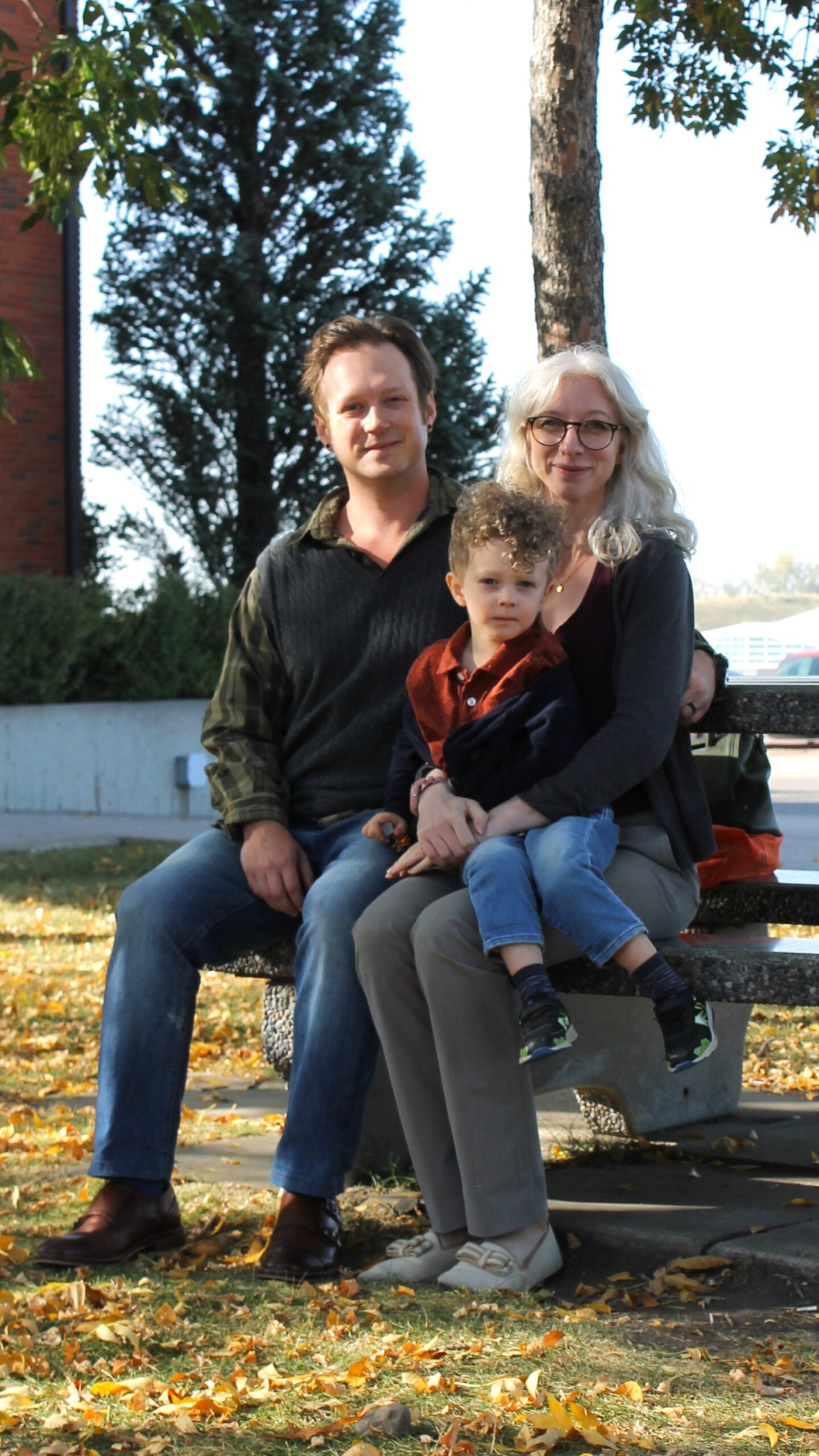 Pamela, Joel and three year old Dima sit on a park bench under large mature trees.