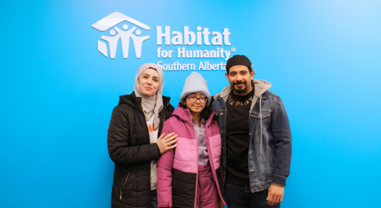 Mohammed, Tiba and their daughter Mayar stand in front of a blue wall with the Habitat for Humanity Southern Alberta logo in white.