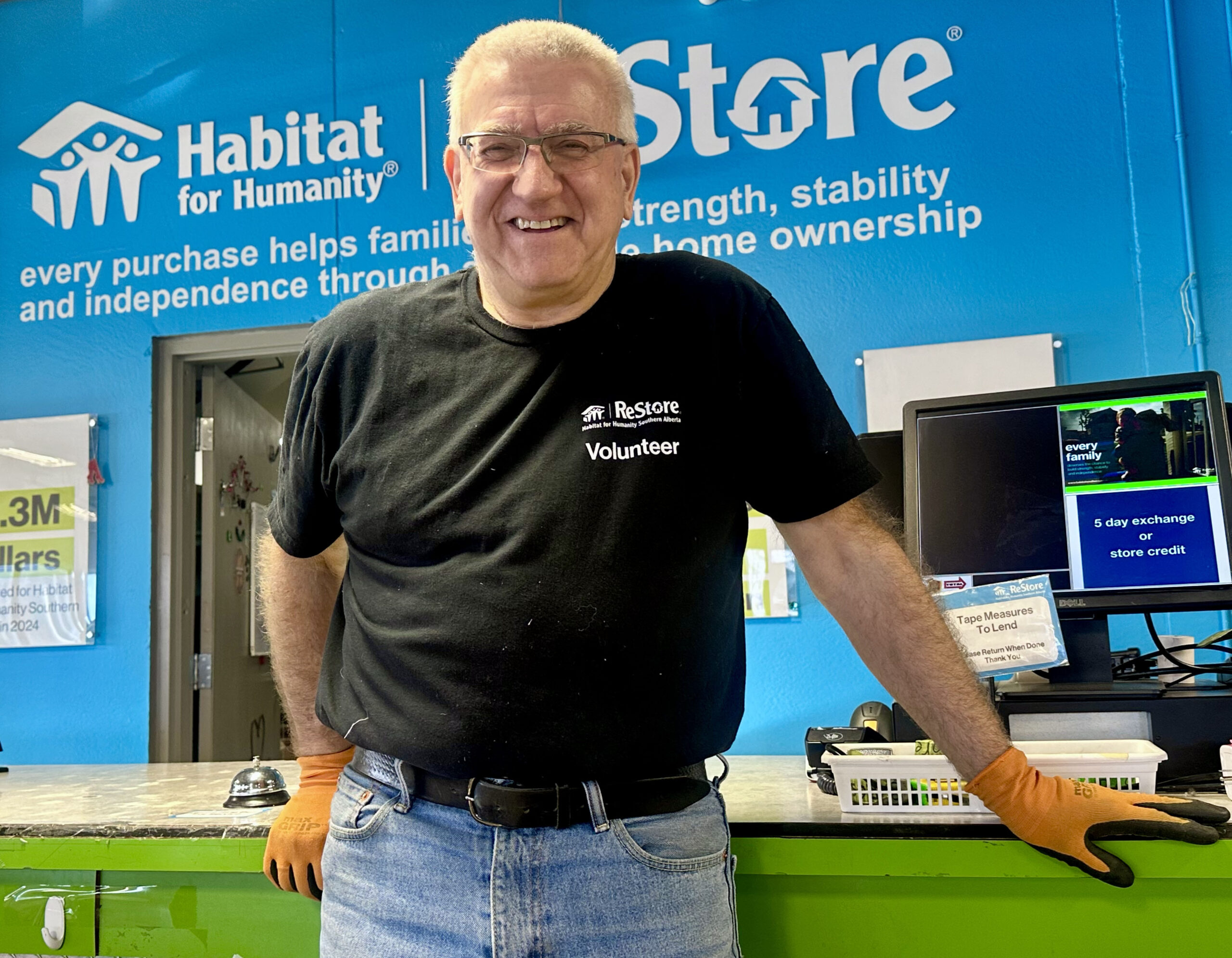 Steve proudly stands in front of the Habitat ReStore blue wall with a large logo wearing a black tshirt with "Volunteer" written on the front.
