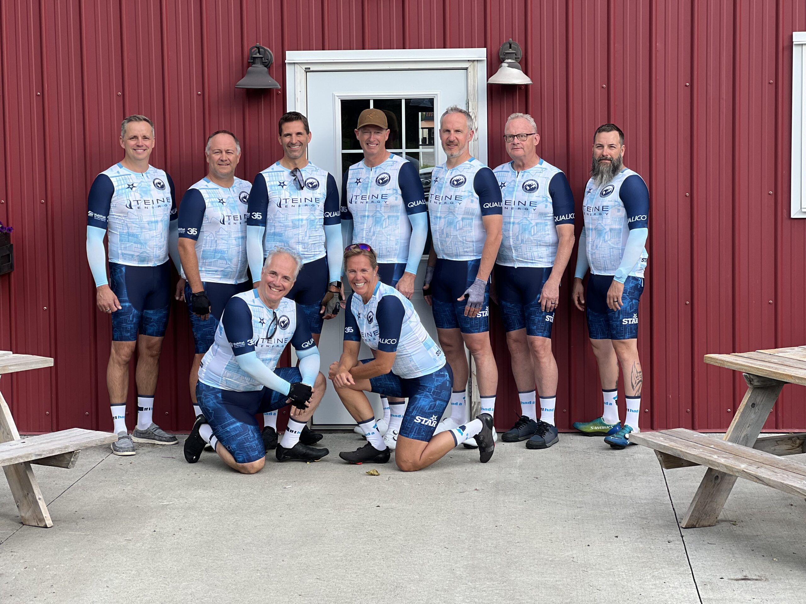 A group of cyclists wearing a light blue kit stand in front of a red wall.