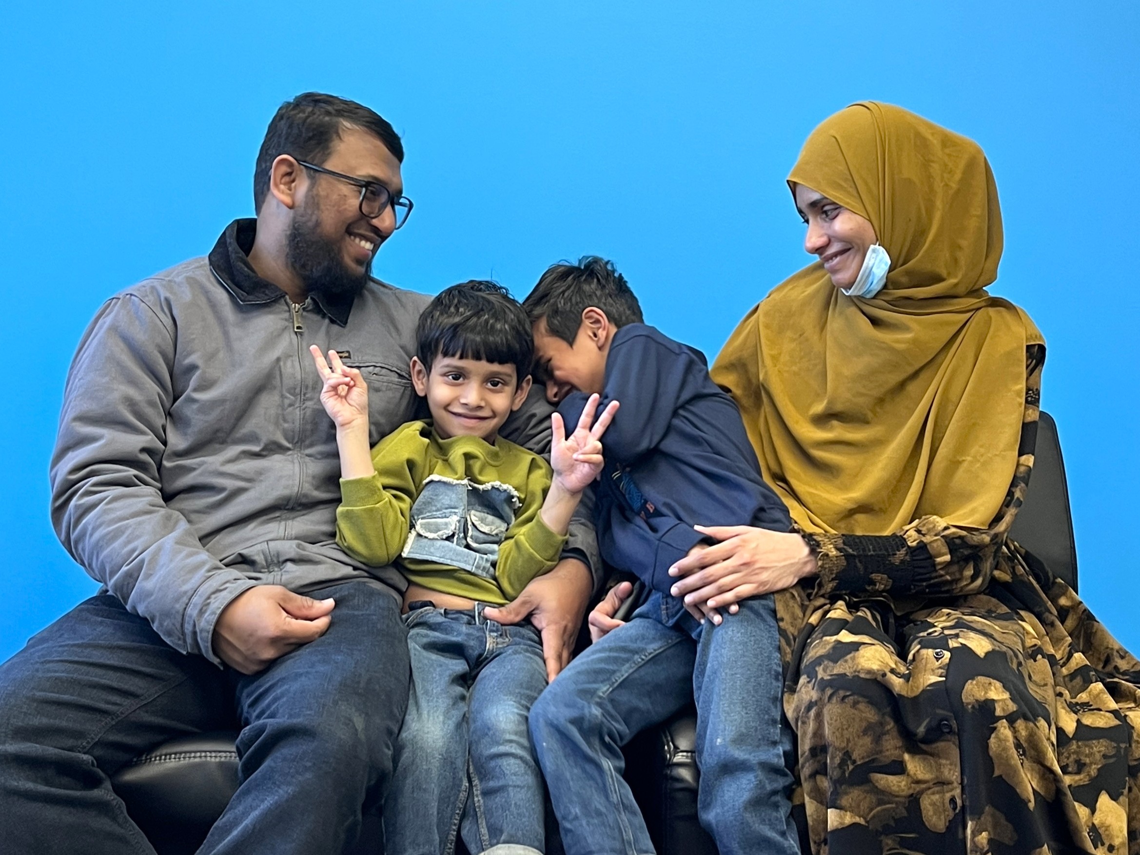 Two parents smile down at their two young boys between them in front of a blue wall.