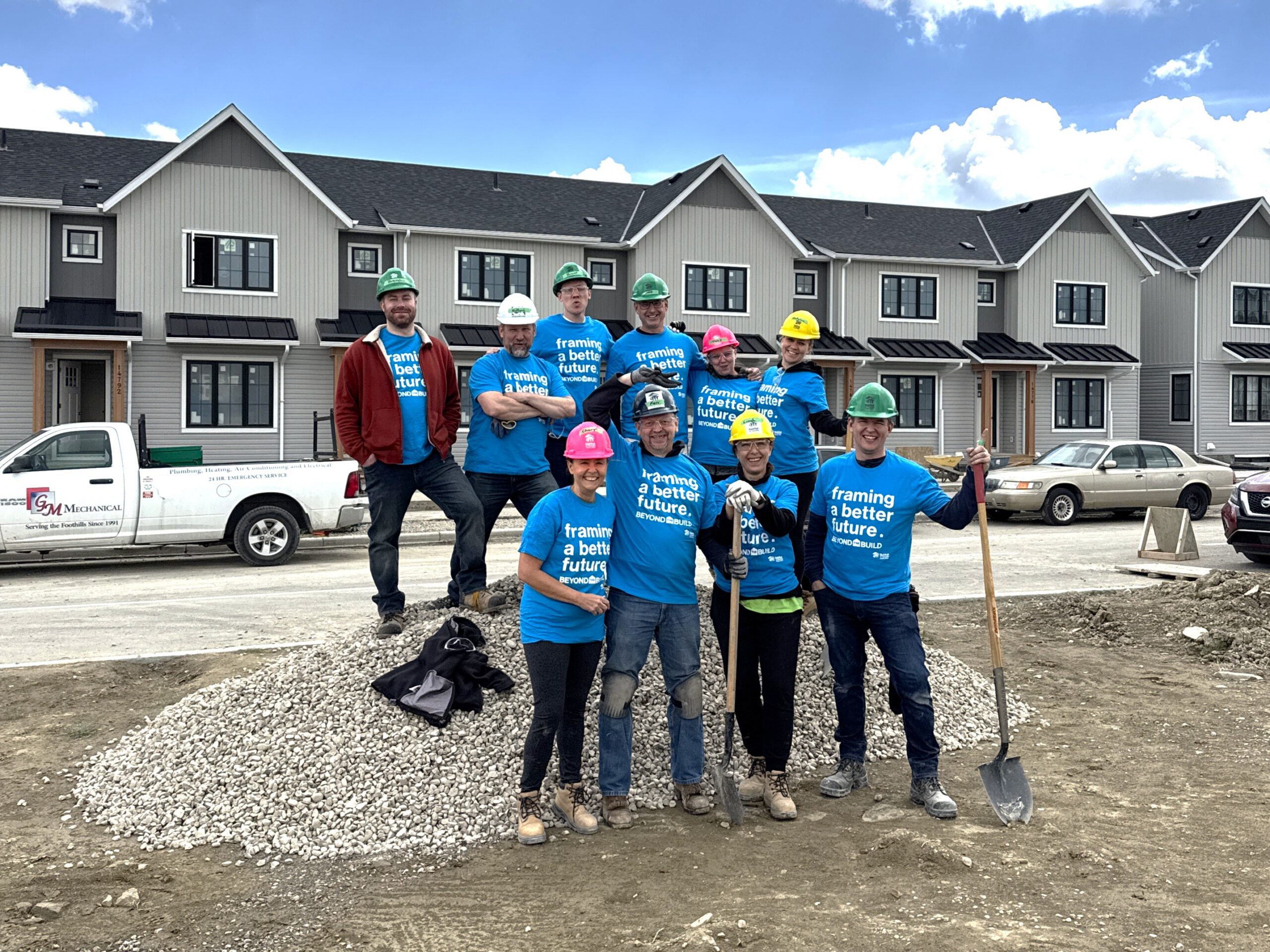 A group of people wearing blue shirts and hardhats stand on top of a rock pile with shovels.
