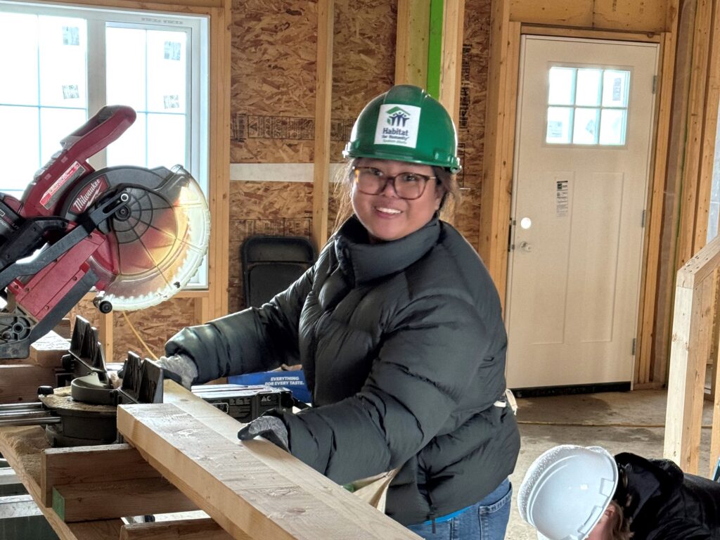 A woman wearing a green hardhat operates a mitre saw on a Habitat build site. 