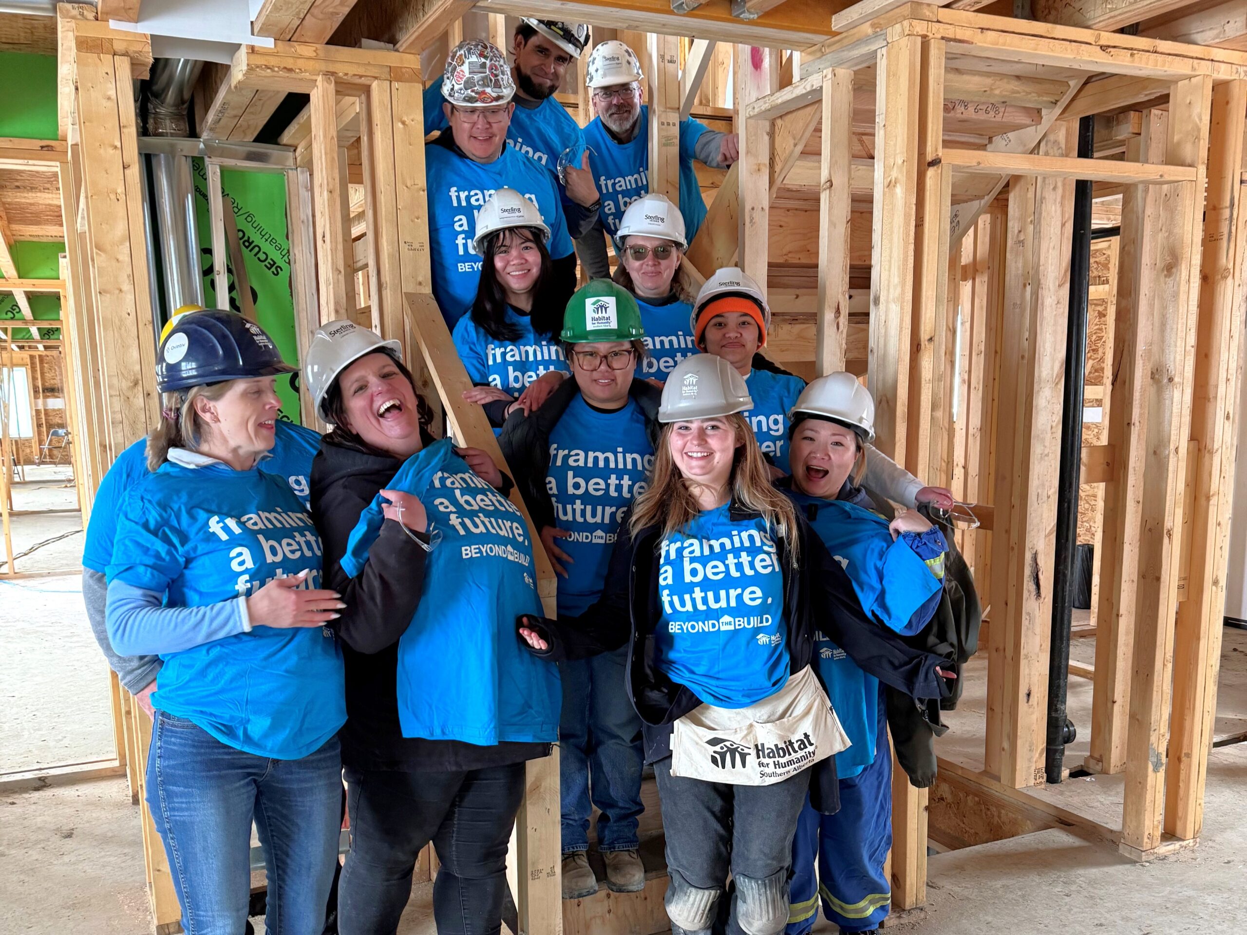 A group of people wearing blue tshirts and hardhats stands in a framed basement.