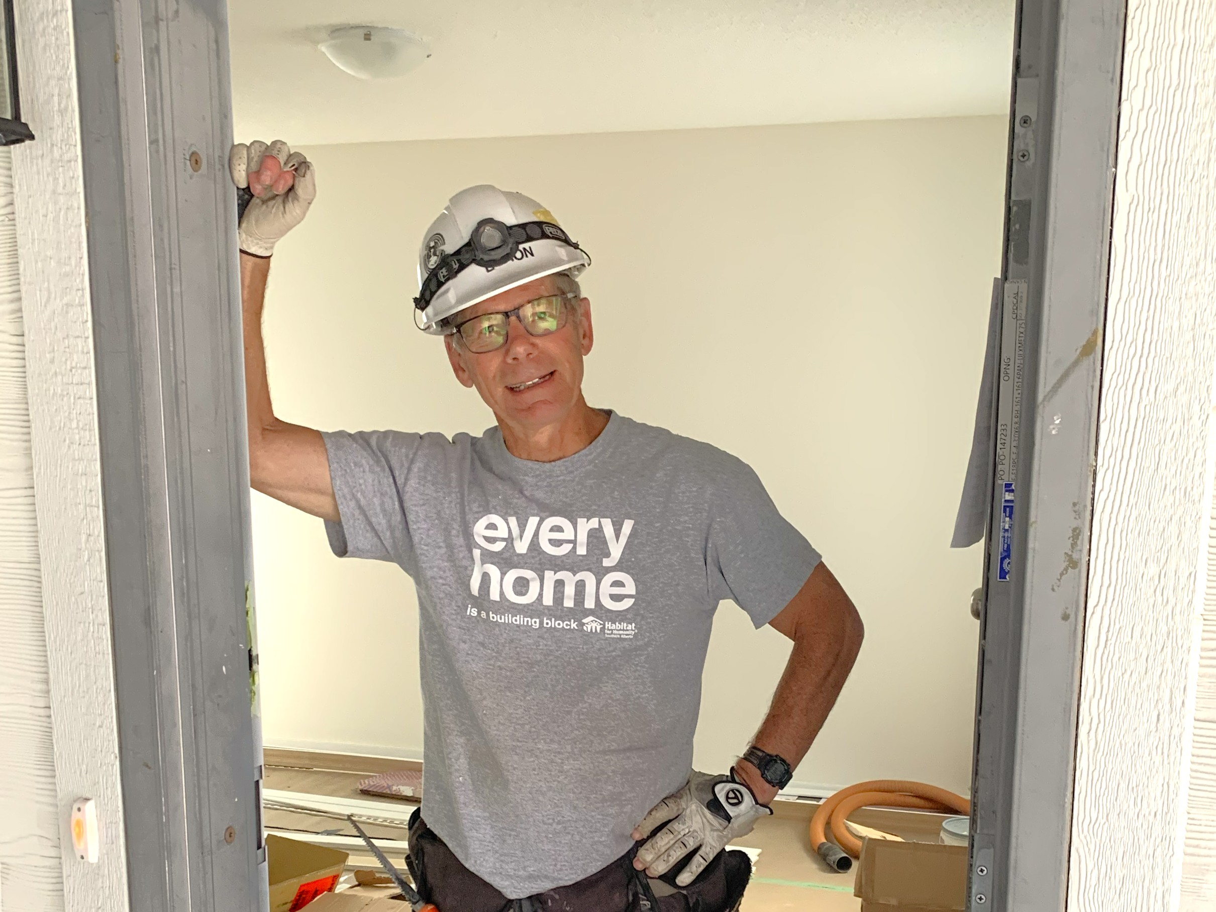 Byron stands in a doorframe wearing a grey 'every home' tshirt and white hardhat.