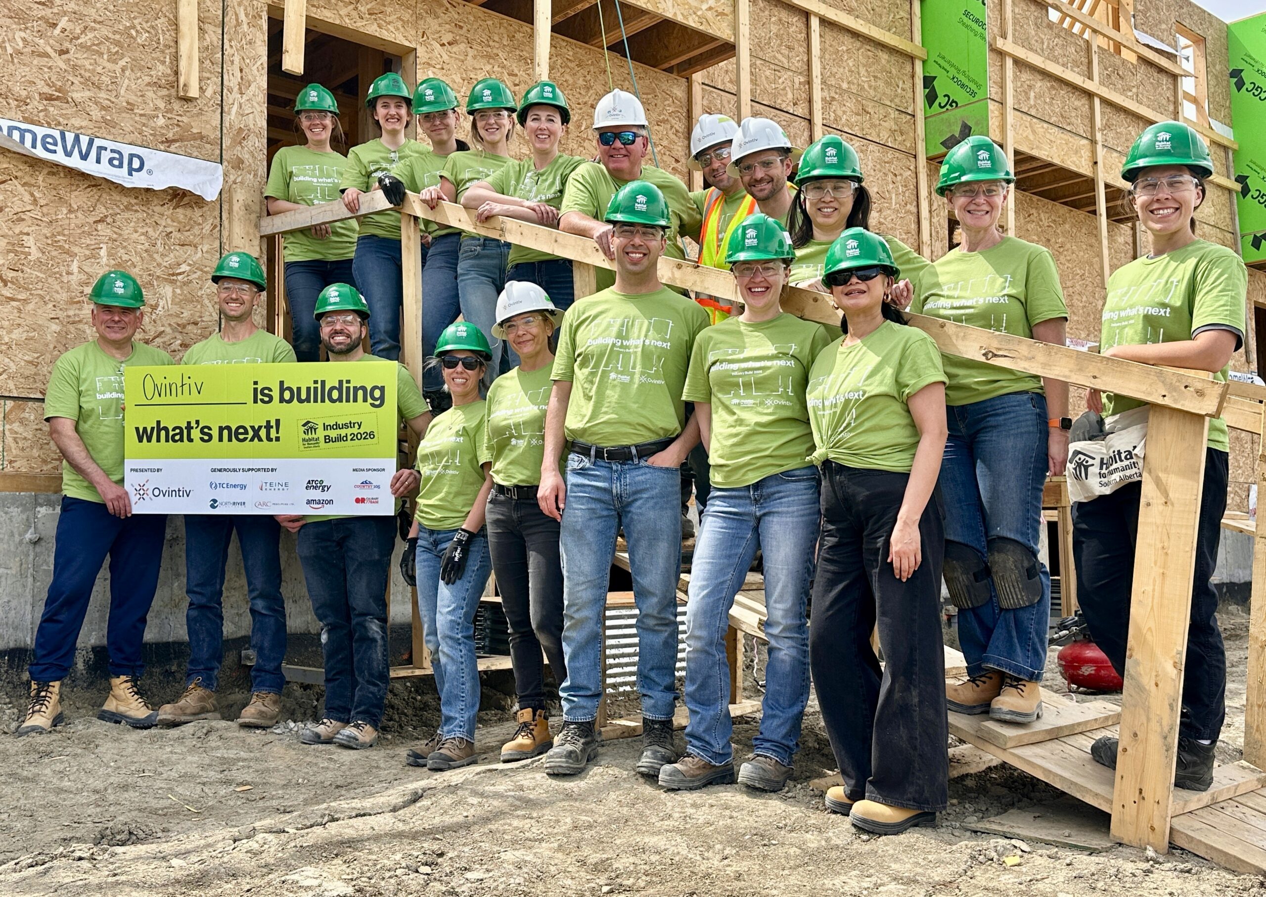 20 volunteers wear green tshirts and hardhats stand smiling in front of an unfinished framed home