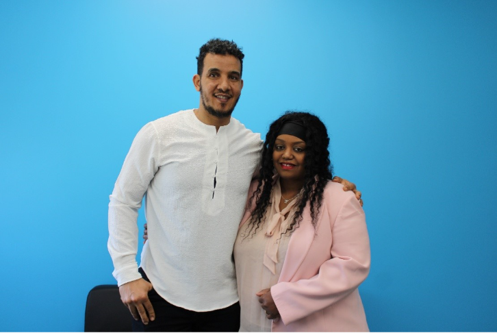 A husband and wife stand together in front of a blue wall.