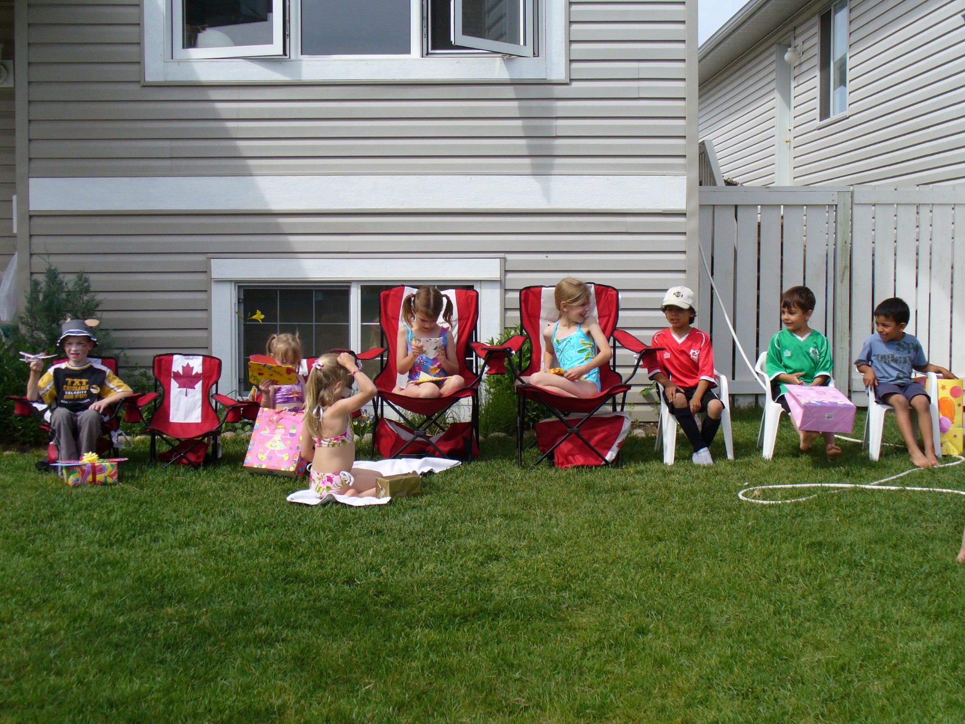 The neighbourhood kids sit on lawn chairs on green grass.