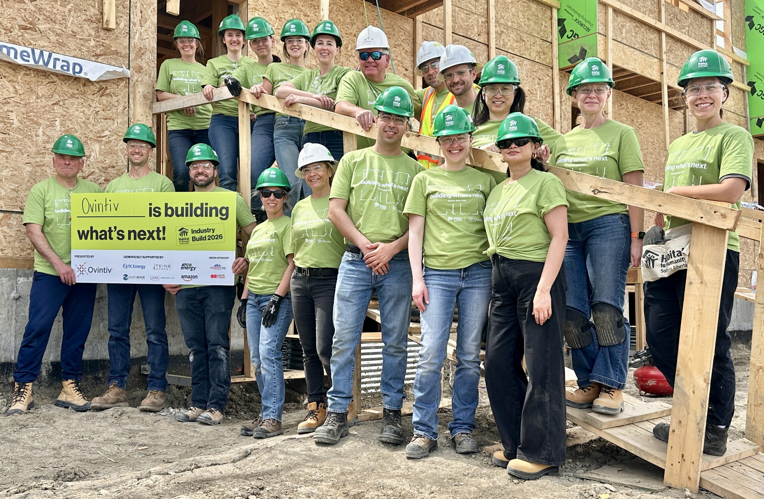 30 men and women wear matching green shirts and stand in front of a home under construction.