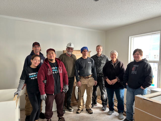 Eight people stand inside an unfinished living room.